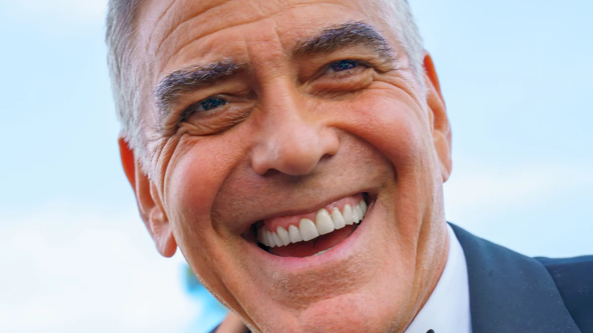 George Clooney sourit en gros plan, vêtu d’un costume sombre et d’une chemise blanche.