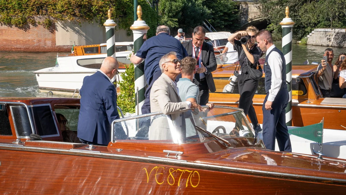 George Clooney, vêtu d’un costume clair et de lunettes de soleil, se tient dans un bateau à moteur en bois entouré de photographes et d’autres embarcations.