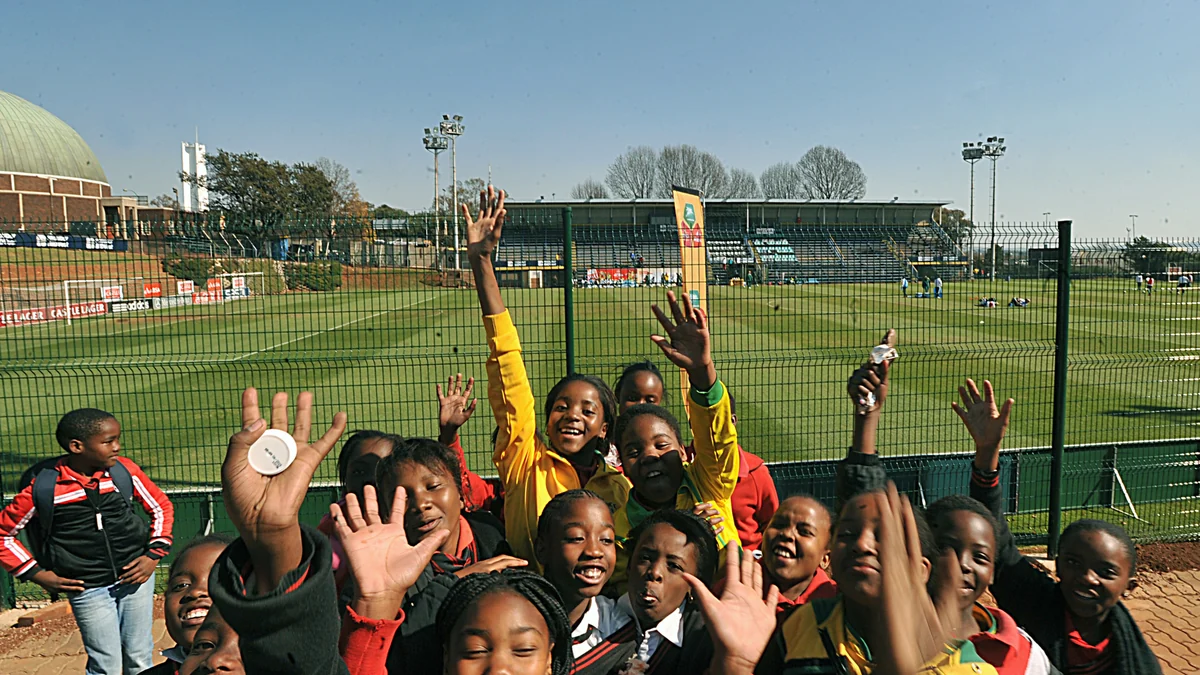 Students wave in front of a field where a football training session is taking place