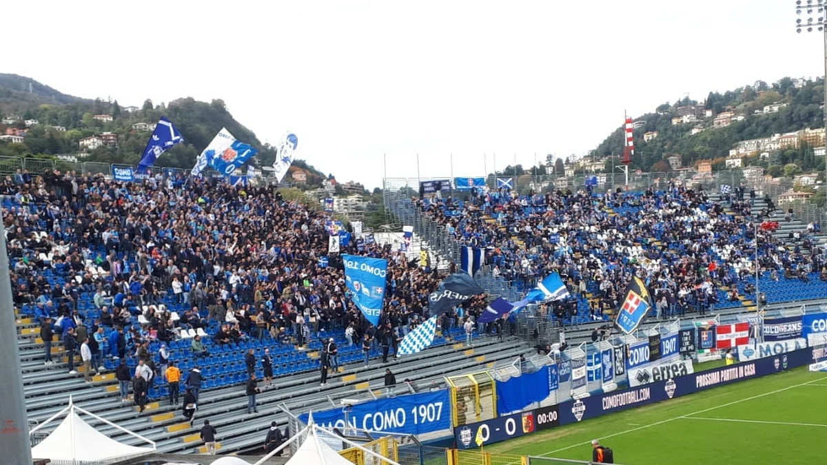 Como supporters with blue flags in an open stand in front of an urban hillside