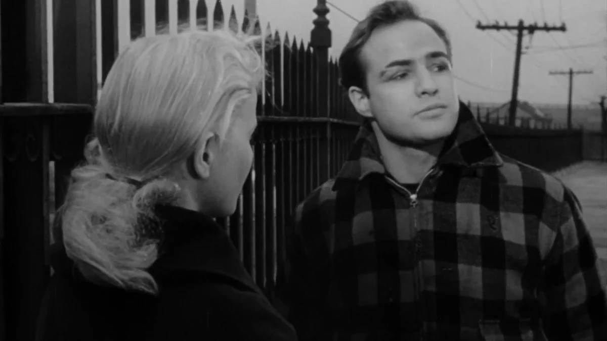 Eva Marie Saint in profile beside Marlon Brando, who wears a checked jacket, in a black-and-white scene