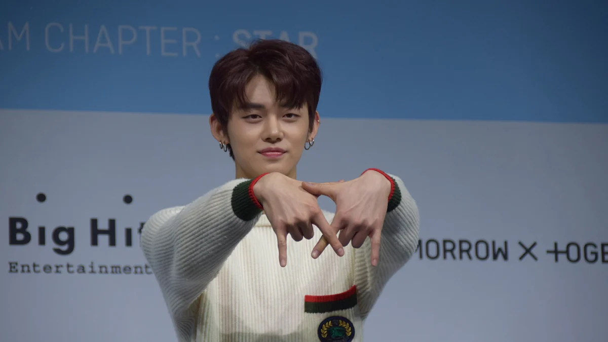 YEONJUN devant un panneau bleu, les bras levés et formant un geste avec les mains