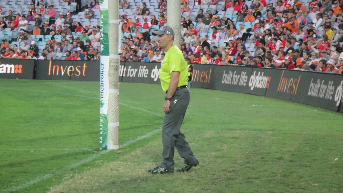 Goal umpire con uniforme verde junto a los postes de un campo, con público al fondo