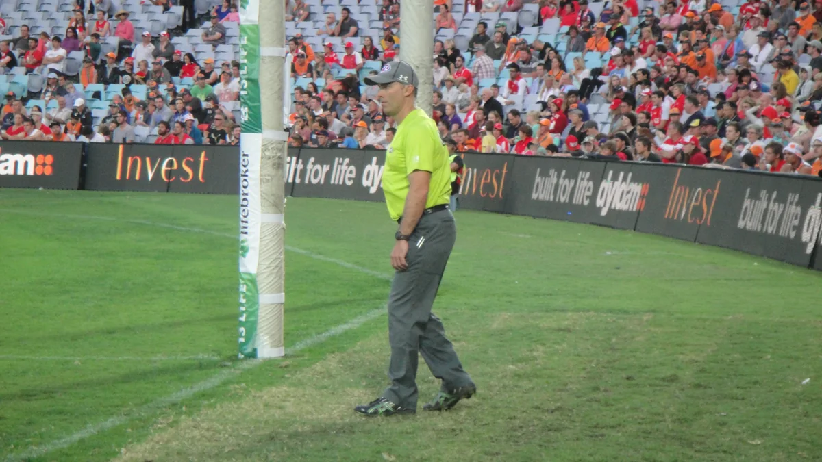 Goal umpire de uniforme verde ao lado dos postes em um campo, com arquibancada ao fundo