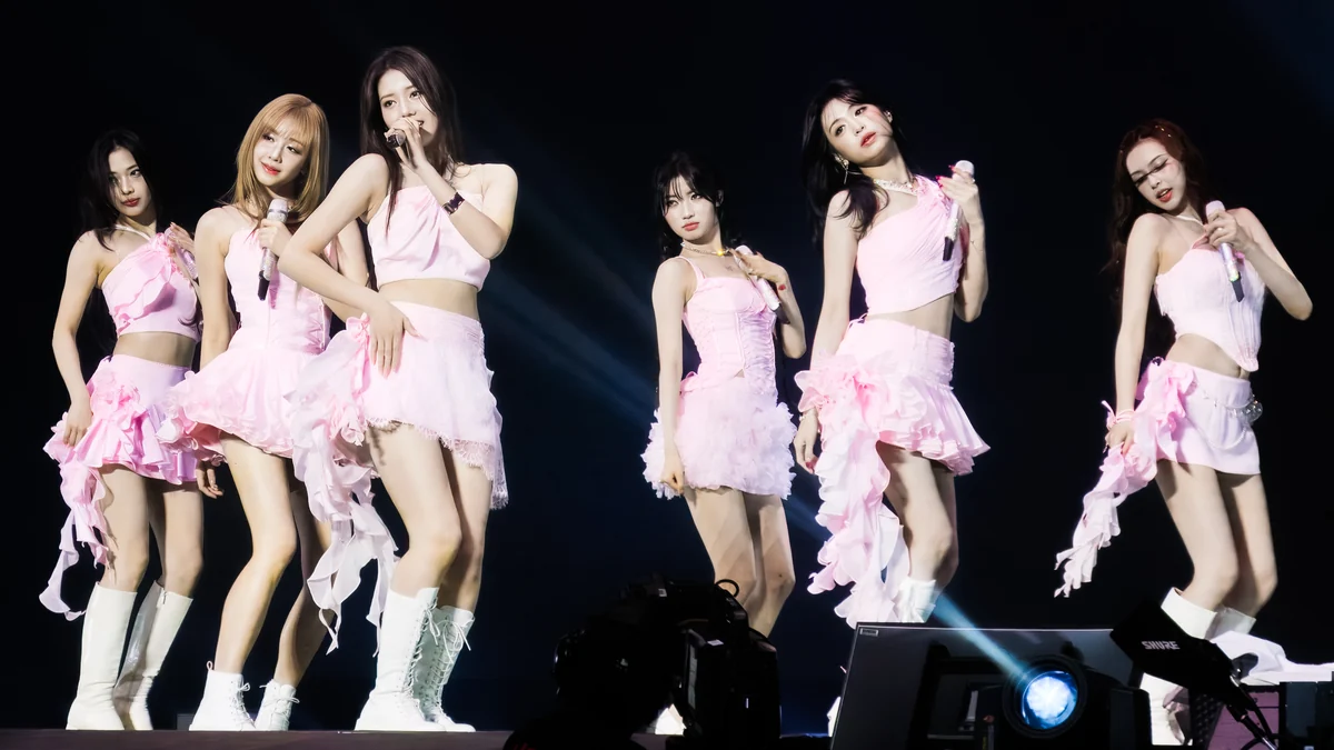 Six BABYMONSTER members perform onstage in pink outfits while holding microphones