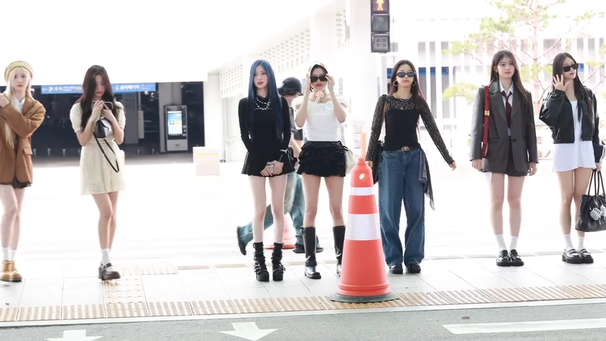 BABYMONSTER members wait beside a pedestrian crossing in an outdoor airport area