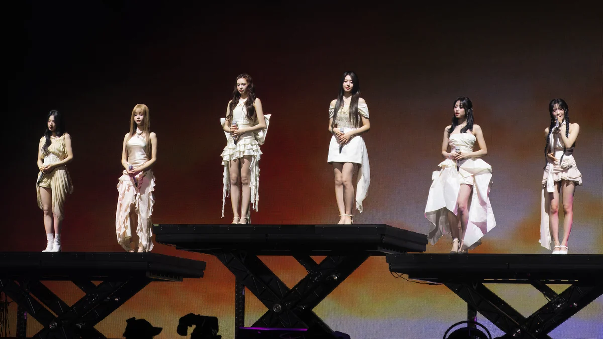 Six BABYMONSTER members stand on raised platforms during a performance with orange lighting