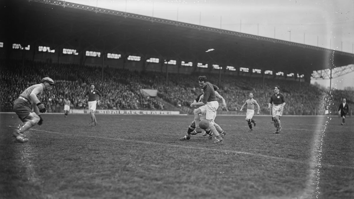 Match de football en noir et blanc avec un attaquant entouré de défenseurs devant une tribune pleine