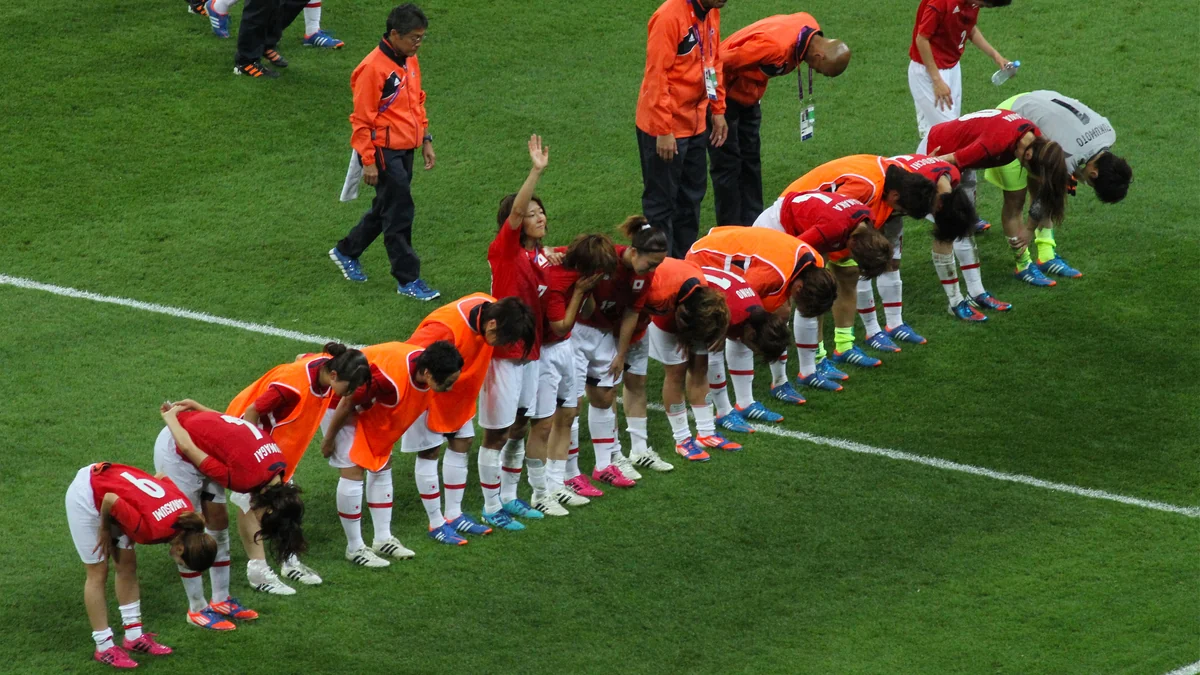 Jugadoras de la selección femenina de Japón, con camisetas rojas, saludan alineadas al público en el campo
