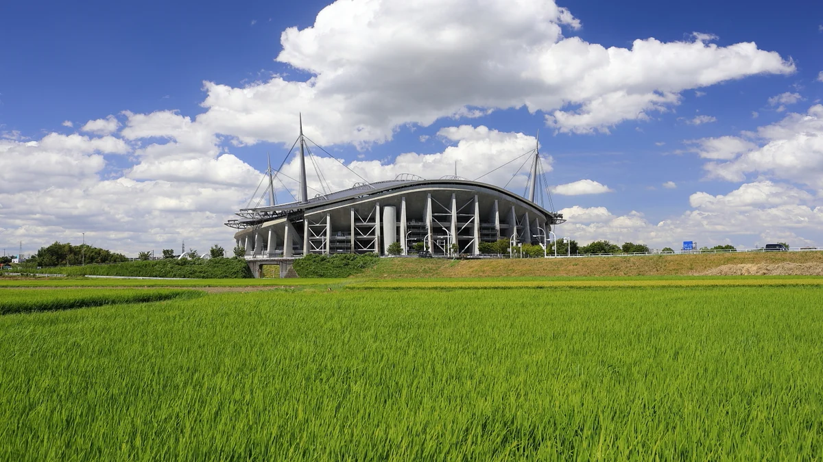 Toyota Stadium visto à distância atrás de um campo verde, sob céu azul com nuvens