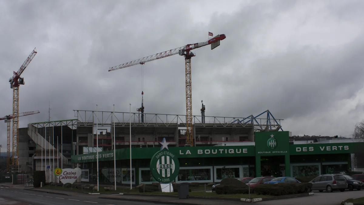 Loja da AS Saint-Étienne diante do estádio, com carros estacionados e guindastes ao fundo