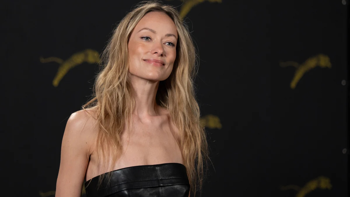 Olivia Wilde pose devant un fond sombre dans une robe noire sans bretelles.