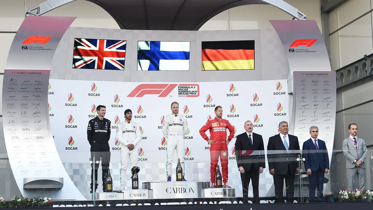 Drivers and officials at a Formula 1 podium ceremony beneath the flags of the United Kingdom, Finland and Germany.