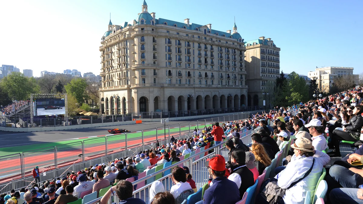 Formula 1 car passes packed grandstands and a large urban building.