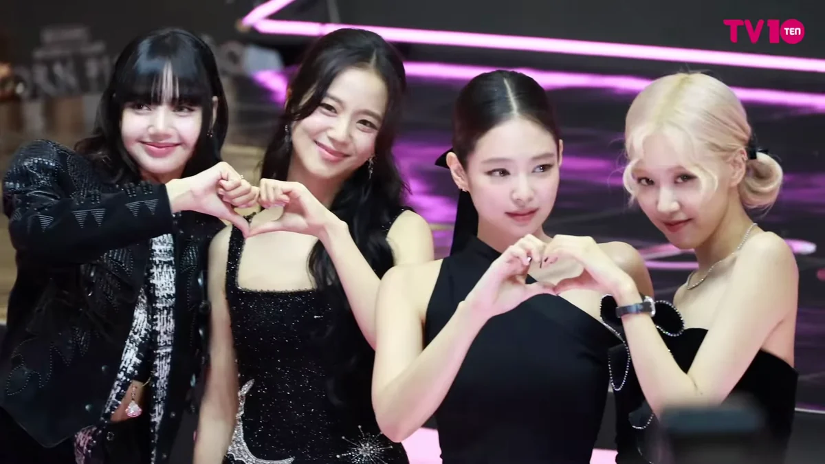 Quatro integrantes do BLACKPINK sorriem e formam dois corações com as mãos diante de luzes rosas.