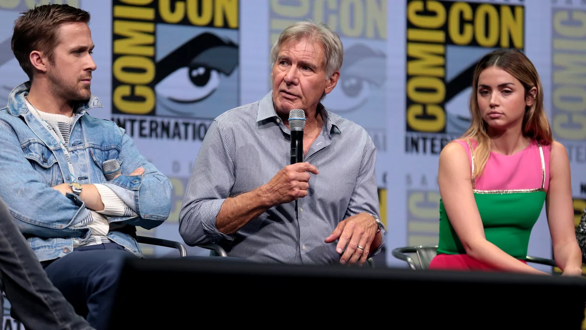 Harrison Ford sostiene un micrófono sentado entre Ryan Gosling y Ana de Armas en un escenario.