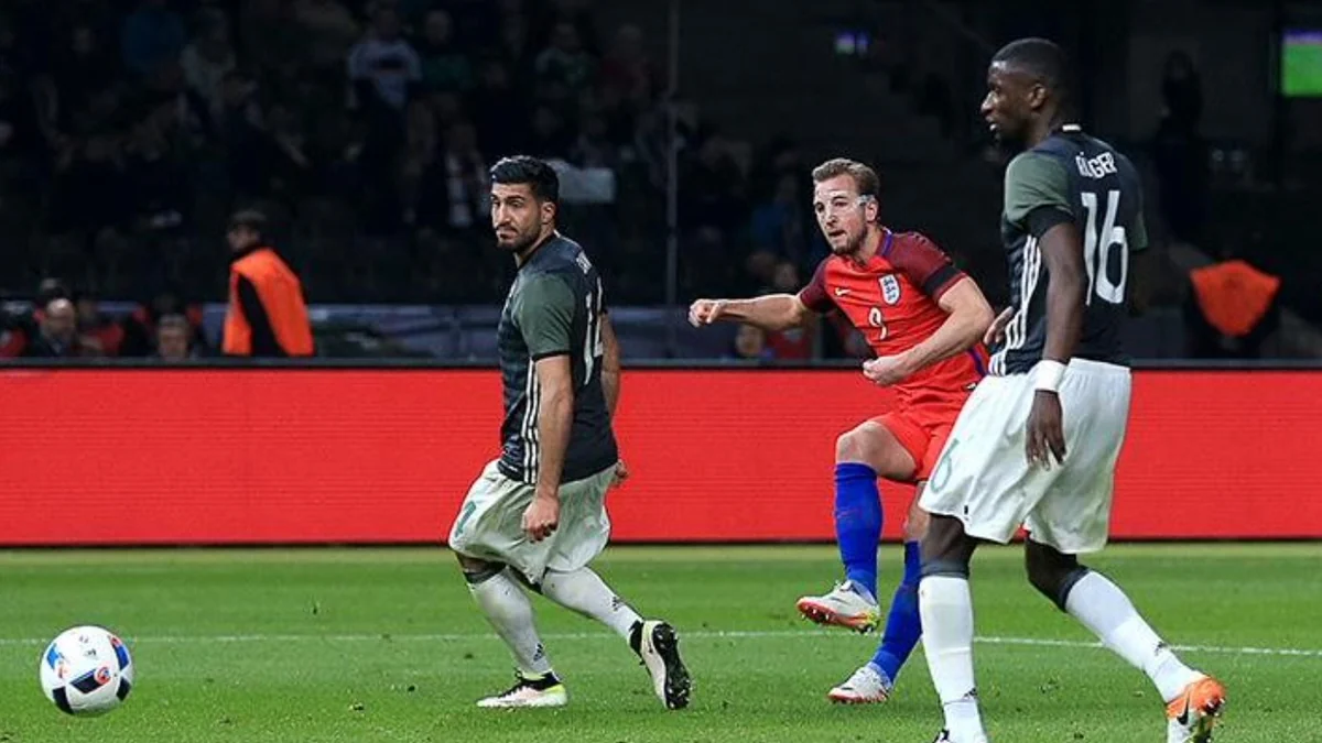 Harry Kane finaliza com a perna direita enquanto dois jogadores da Alemanha acompanham a jogada.