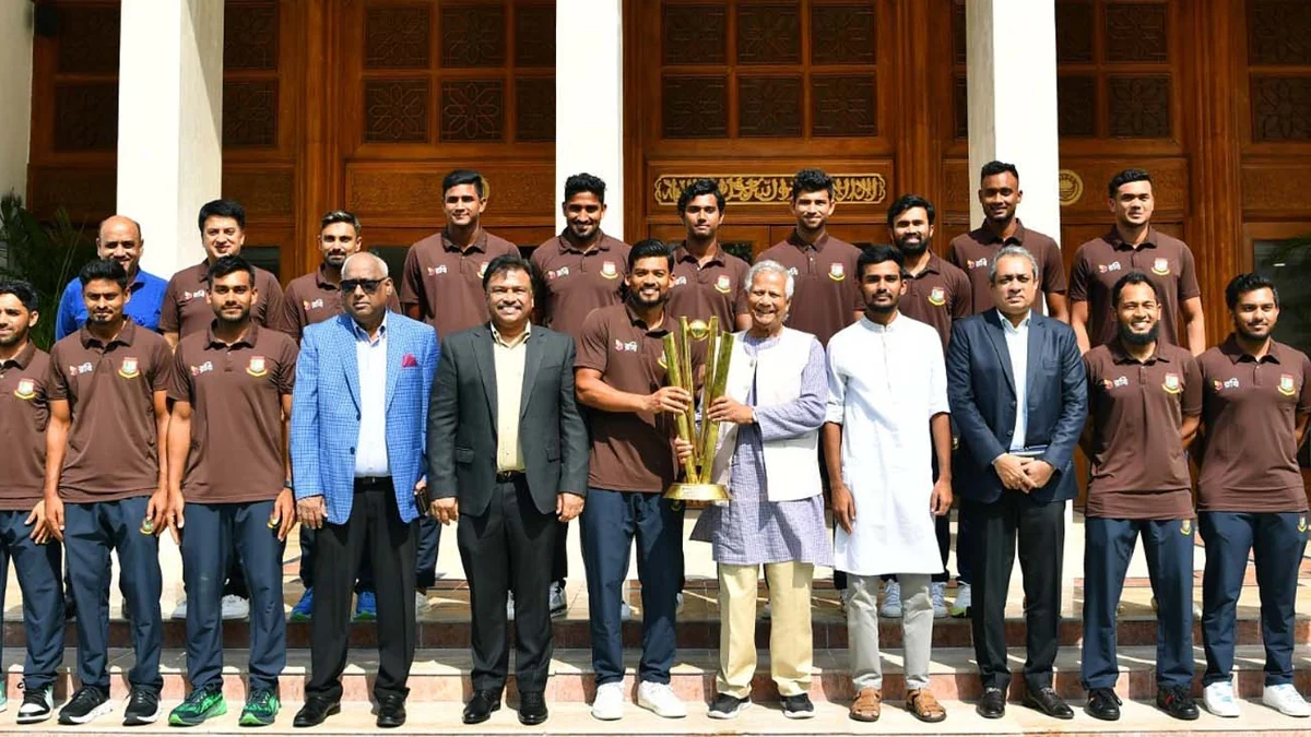 Grupo de jogadores e dirigentes de Bangladesh posa nos degraus de um edifício, com um troféu dourado ao centro.