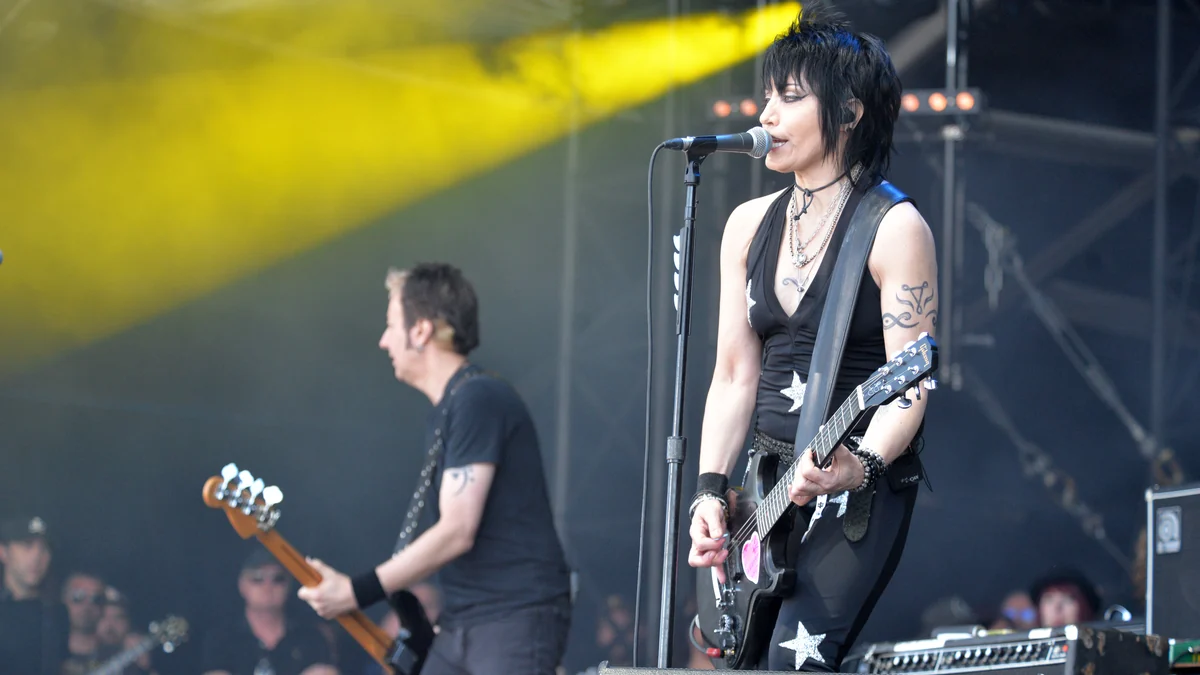 Joan Jett canta e toca guitarra no palco, com um baixista ao fundo e feixe de luz amarela