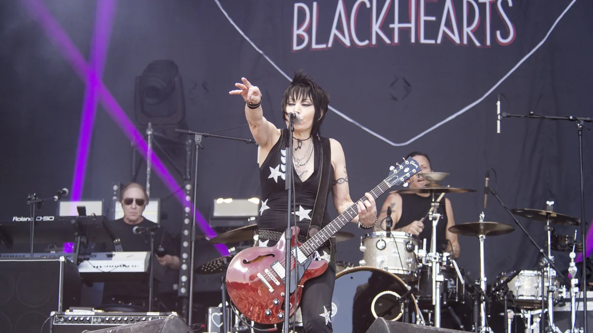 Joan Jett toca guitarra e estende o braço diante da banda em um palco ao ar livre