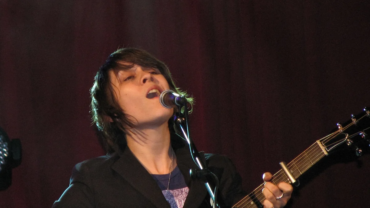 Tegan canta ao microfone enquanto toca guitarra em um palco