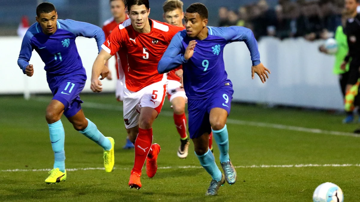 Three young footballers chase the ball, two wearing blue and one wearing red