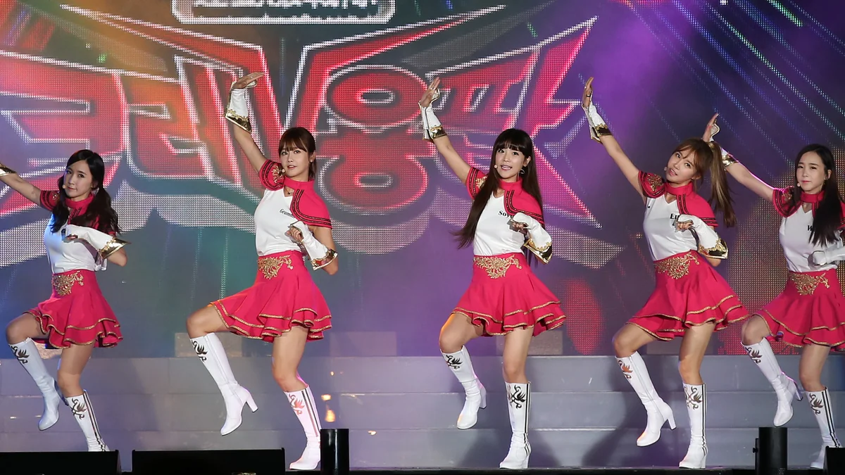 Cinco integrantes de um grupo de K-pop dançam no palco com figurinos rosa e branco.