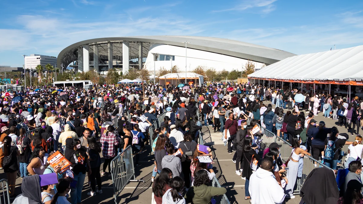 Vista amplia de una multitud en pasillos de vallas, con un estadio y una carpa blanca al fondo