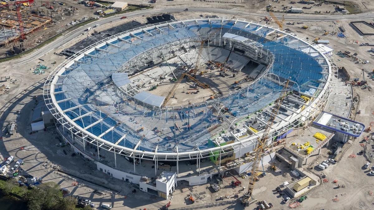 Vista aérea de un estadio ovalado en construcción, rodeado de grúas y zonas de obra