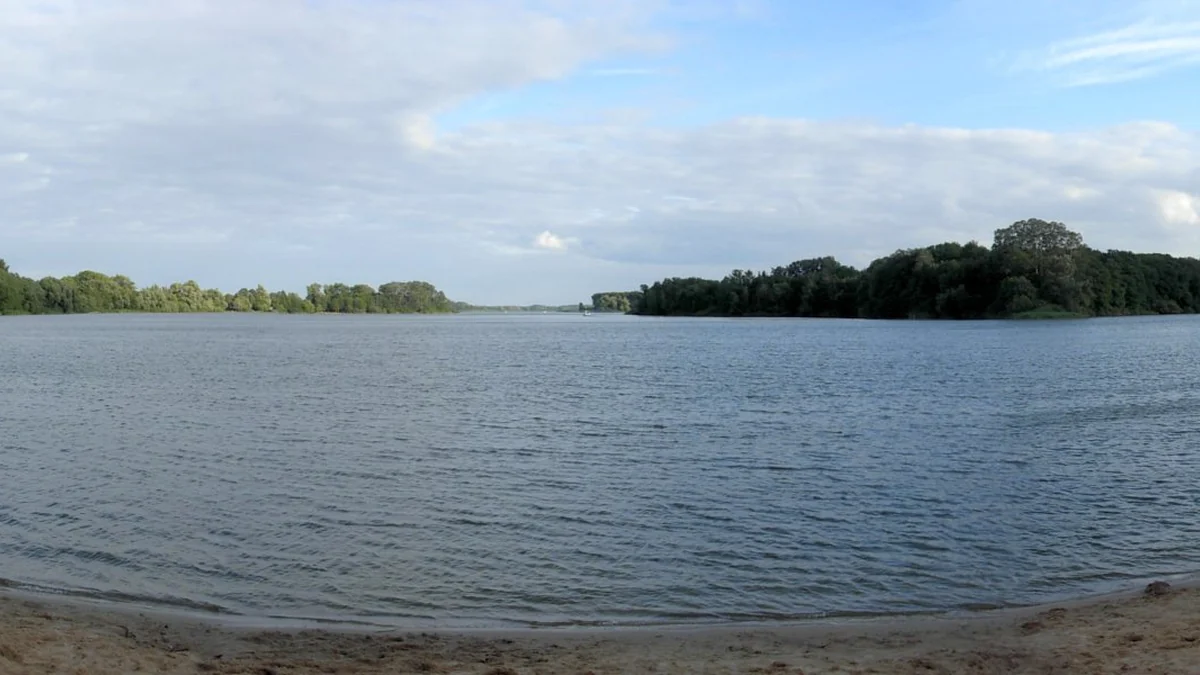 Vaste lac vu depuis une rive sablonneuse, avec des îles boisées au loin et un ciel partiellement nuageux.