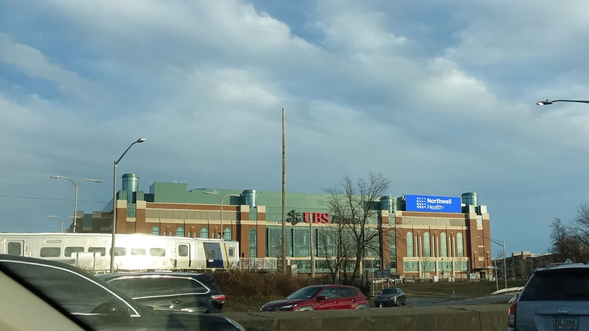 Exterior view of UBS Arena with cars and a train in the foreground beneath cloudy skies