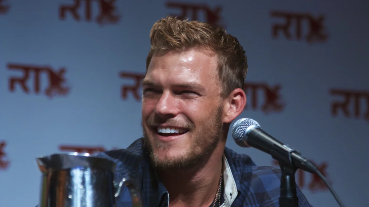 Alan Ritchson sorri diante de um microfone durante um painel