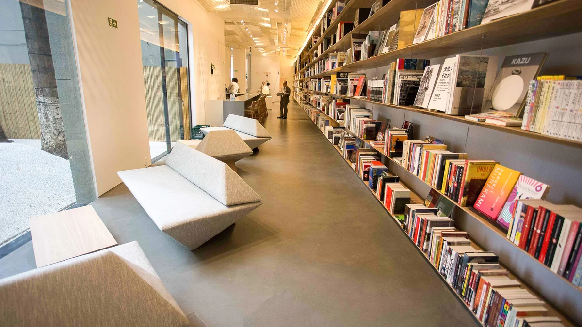 Corredor interno com estantes cheias de livros à direita, bancos claros e janelas à esquerda.