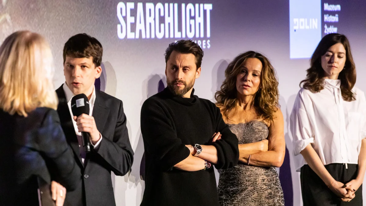 Jesse Eisenberg fala ao microfone em um palco, ao lado de três participantes