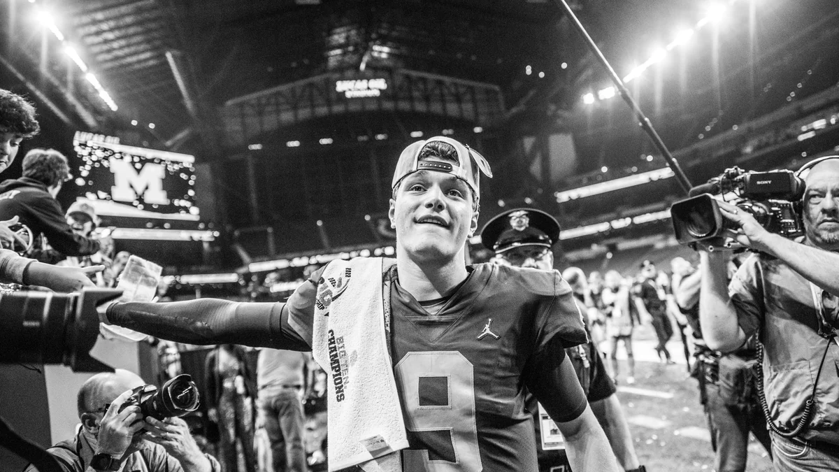 J.J. McCarthy con uniforme de Michigan y gorra hacia atrás, rodeado de fotógrafos en el campo