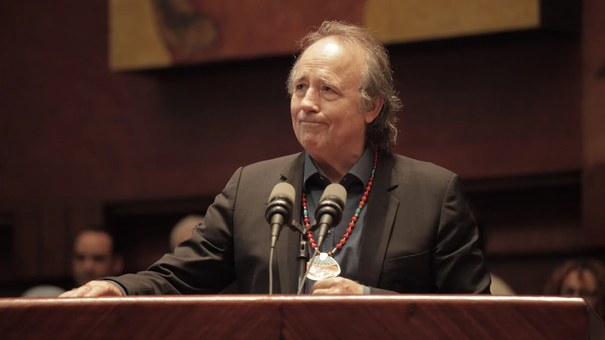 Joan Manuel Serrat standing at a lectern behind two microphones