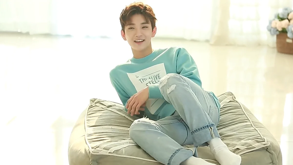 Joshua smiles while seated on a cushion in a light-blue sweatshirt, jeans and white socks