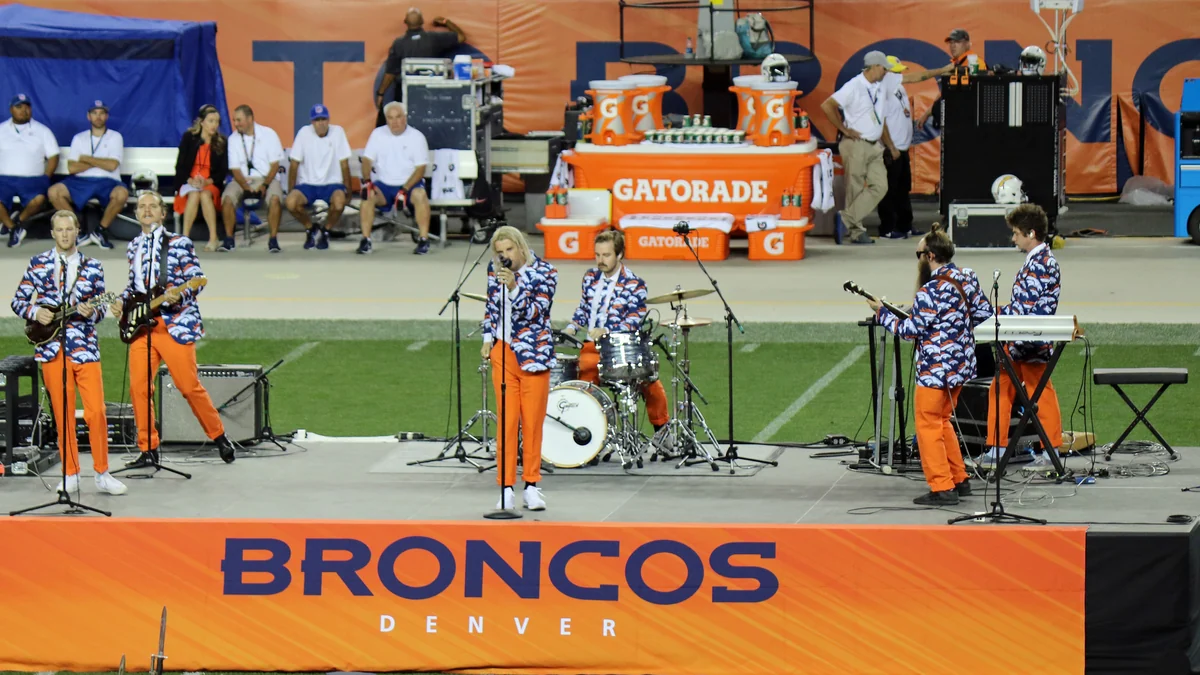 Un groupe se produit sur un terrain de football devant une bannière orange des Denver Broncos