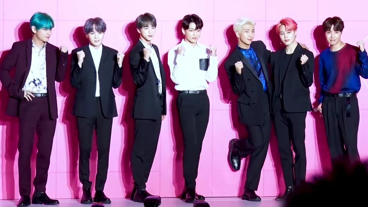 All seven BTS members pose with raised fists against a pink wall.
