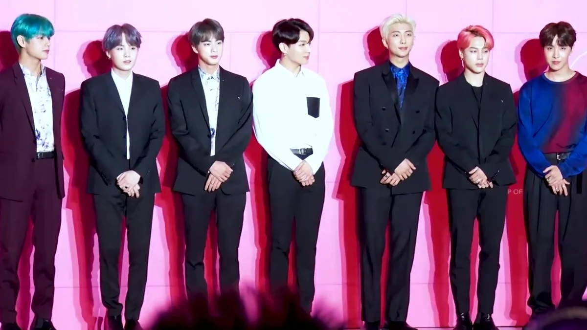 All seven BTS members stand in a line against a pink wall during a press conference.