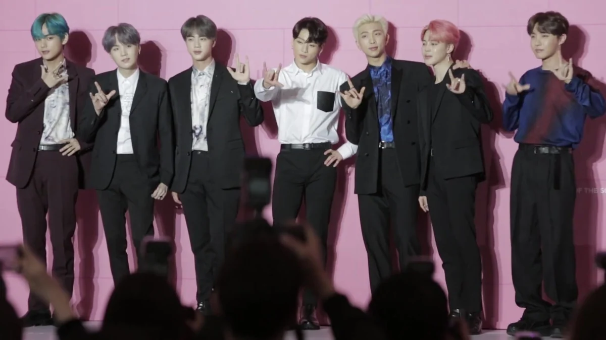 All seven BTS members pose onstage against a pink wall, with audience cameras in the foreground.
