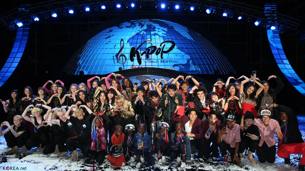 Grande grupo de participantes posa no palco sob o letreiro K-pop World Festival