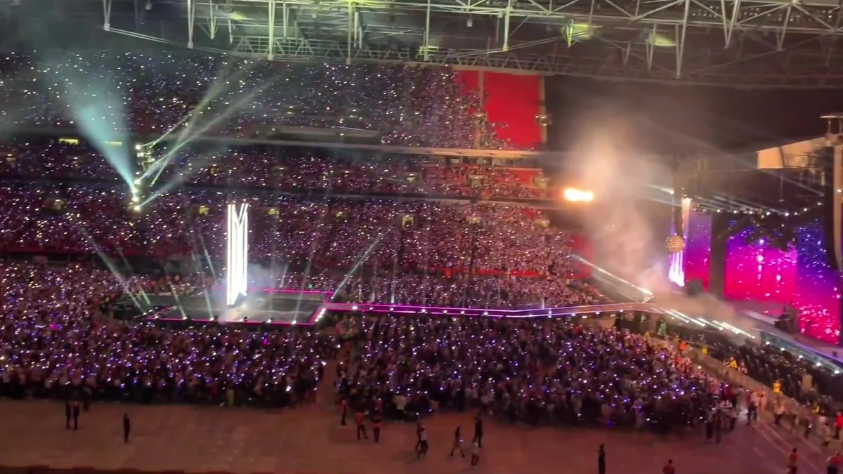 Packed stadium with a pink-lit stage, light beams and an audience holding light sticks