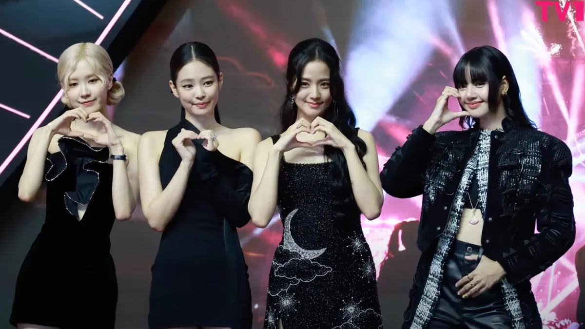 Four BLACKPINK members make heart gestures in front of an illuminated backdrop.