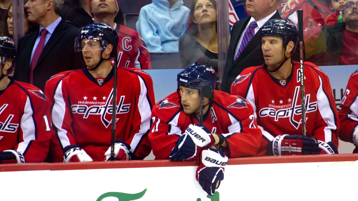 Três jogadores do Washington Capitals com camisas vermelhas sentados no banco da equipe.