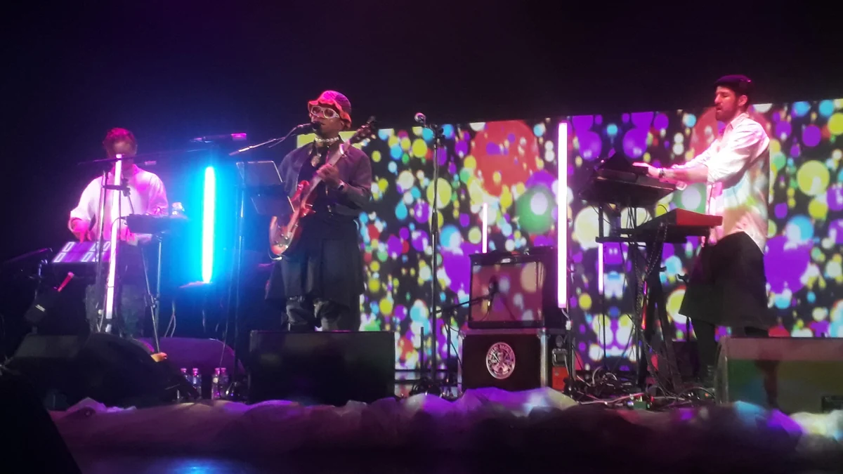 Blick Bassy toca guitarra e canta no centro de um palco com músicos e painéis coloridos