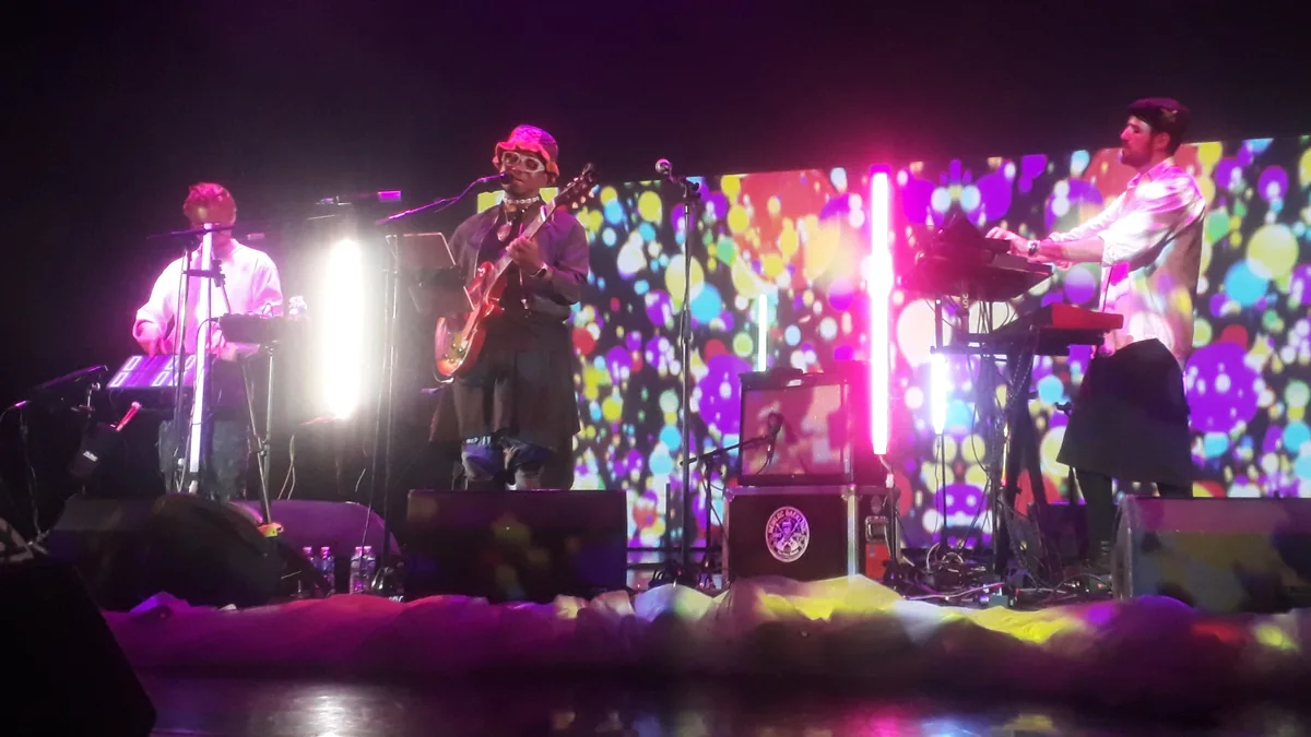Blick Bassy sings and plays guitar between two musicians on a brightly lit stage