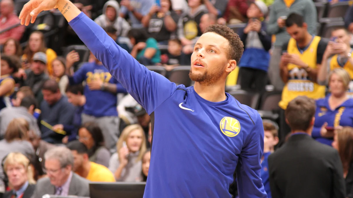 Stephen Curry, con sudadera azul de los Warriors, levanta el brazo ante la grada.