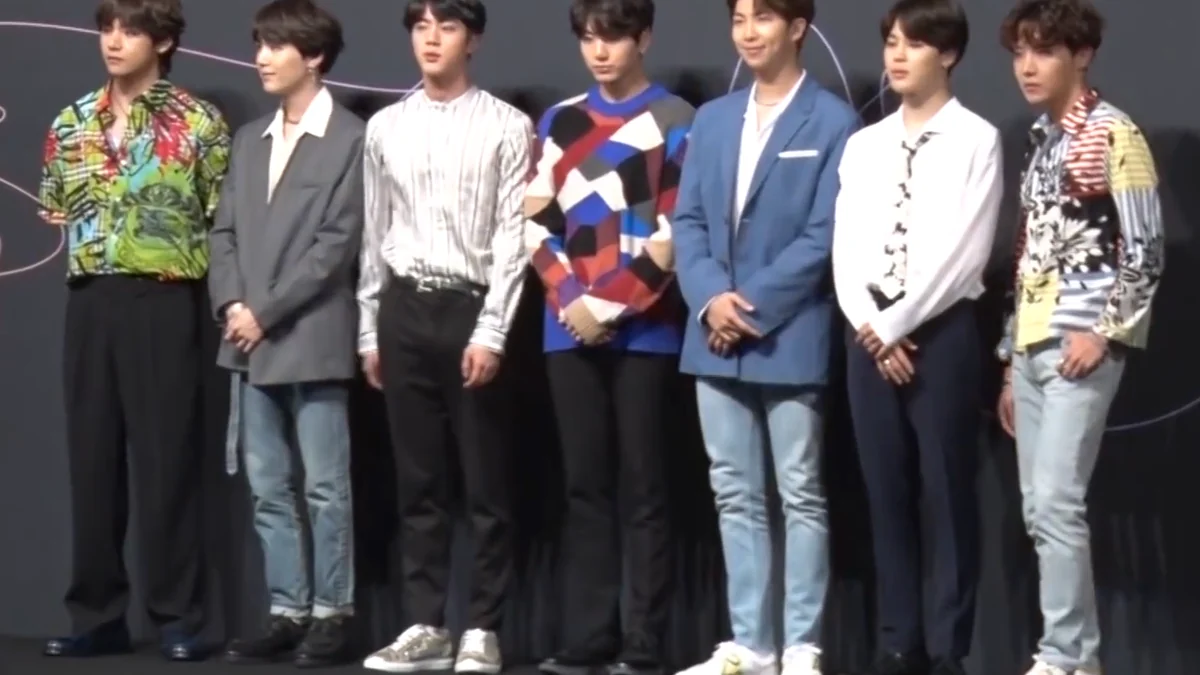 Los siete integrantes de BTS están de pie, uno junto a otro, con ropa colorida y formal sobre un escenario oscuro