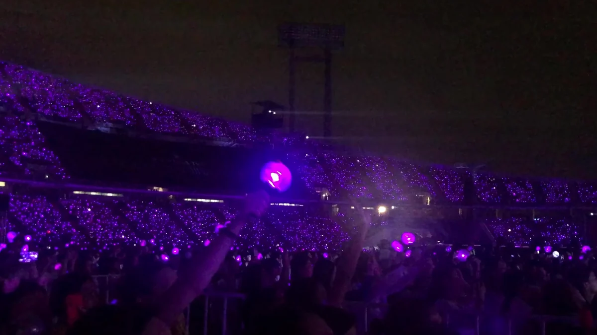 A large crowd fills an arena illuminated by thousands of purple light sticks.
